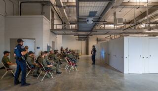 A group of uniformed individuals sits in chairs, while two officers stand and converse in a spacious, industrial setting.