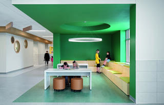 A modern, vibrant space with green walls, children engaged in activities, and a cozy seating area featuring round tables and casual seating.