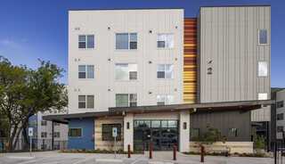 A modern multi-story apartment building features a mix of colorful and neutral facades, with a welcoming entrance and landscaped surroundings.