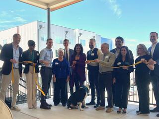 A group of people, including a dog, gathers with a cut yellow ribbon for a ceremonial opening, showcasing a mix of business attire and casual wear.