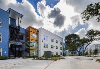 Colorful modern apartments with varying architectural designs under a dramatic sky, surrounded by green trees and open spaces.