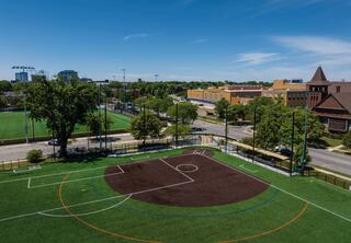 A well-maintained soccer field with vibrant artificial turf is surrounded by trees and urban buildings under a clear blue sky.