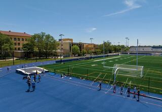A vibrant sports field with a blue track and goalposts, surrounded by greenery and school buildings, filled with active athletes.