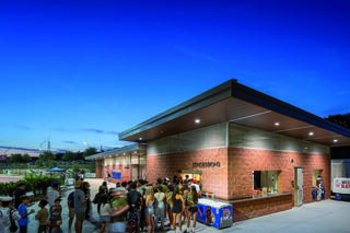 A crowd gathers outside a modern concession stand at dusk, awaiting food and drinks, illuminated by bright lights and a clear evening sky.