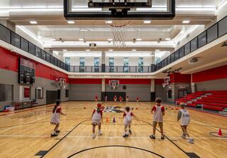 A spacious gymnasium with hardwood floors, multiple basketball hoops, and children practicing drills, surrounded by vibrant red and gray walls.