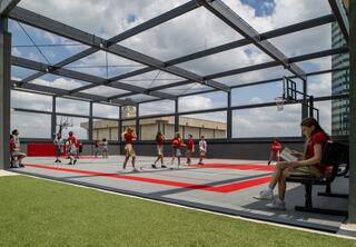 A semi-outdoor basketball court hosts a lively game, with players in red jerseys and a girl sitting nearby, enjoying the view.