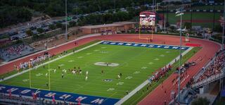 Aerial view of a vibrant football game in a well-lit stadium, with crowds cheering and a digital scoreboard in the background.