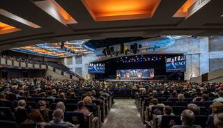 A large audience fills a modern auditorium, facing a brightly lit stage adorned with screens and banners, creating an engaging atmosphere.