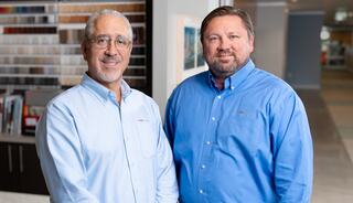 Two men in blue dress shirts pose together in an office setting, smiling confidently with colorful design samples in the background.