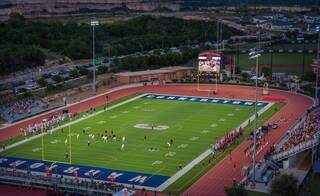 A vibrant football field hosts a game under stadium lights, surrounded by spectators and picturesque views of the landscape beyond.