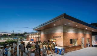 A crowd gathers outside a concession stand at dusk, with bright lights illuminating the building and a lively atmosphere.