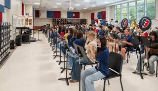 A large group of students participates in a music rehearsal, playing various instruments in a brightly lit classroom setting.