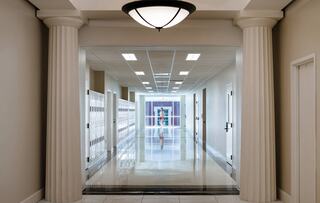A spacious commercial corridor with glossy floors, columns, lockers on one side, and a bright, inviting atmosphere leading to a distant exit.