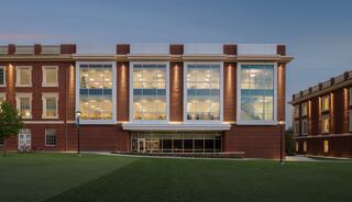 A modern, glass-fronted building is framed by manicured lawns and trees, showcasing bright interior lights against a dusk sky.