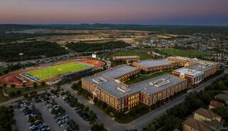 Aerial view of a modern school complex with sports fields and parking, surrounded by greenery and hills at dusk.