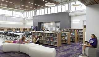 A modern library features students reading on furniture, surrounded by shelves of books, with a spacious, well-lit design.