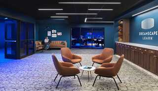 A modern lounge area features stylish orange chairs, a cozy couch, and a blue-lit ambiance, promoting a creative learning environment.