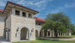 A modern stone building featuring arched entrances, a red-tiled roof, and lush greenery surrounding a central tree and lawn area.