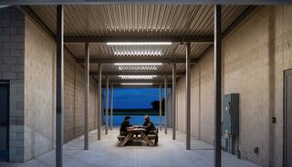 Two people sit at a picnic table in a spacious, industrial-style area, surrounded by concrete walls and illuminated by overhead lights.