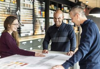 Three people are discussing plans at a table, smiling and engaged, with color samples displayed on shelves behind them.