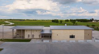 A modern, one-story building with a metal roof, surrounded by green fields and a winding path under a partly cloudy sky.