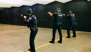 Three police officers are positioned in a training facility, aiming their rifles toward a target area, focused on marksmanship skills.