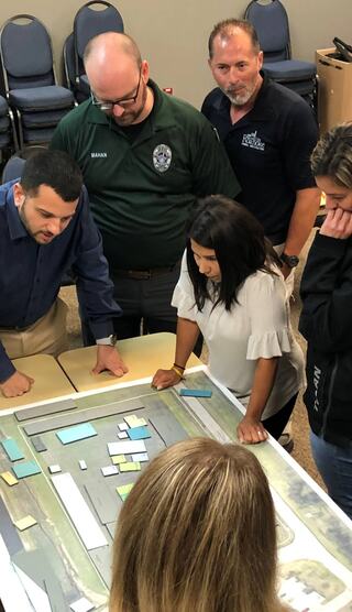 A group of people collaborates around a table, studying a large map with colored pieces, engaged in discussion and planning.