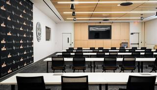 A modern conference room features rows of black chairs and tables, a screen on a wooden wall, and branding on the back wall.