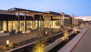 A modern building with a sleek design, featuring large glass windows and landscaped pathways, illuminated softly at dusk.