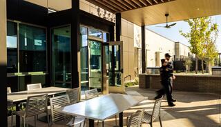 A modern outdoor space features a table and chairs, with a glass entrance nearby. A person walks by, framed by a sunny backdrop.