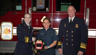 Three firefighters stand together in front of a fire truck, exchanging a helmet in a moment of celebration and camaraderie.