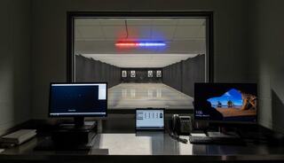 A shooting range view through a glass window, featuring targets, two monitors, and red and blue lights above, indicating an active setup.