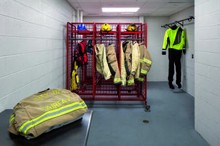 A locker room displays various firefighting gear, including jackets, helmets, and a high-visibility suit, organized on racks and hooks.