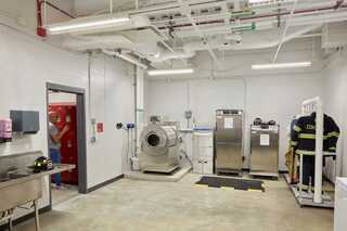A clean, industrial laundry room features stainless steel machines, a sink, storage space, and a uniform on a stand.