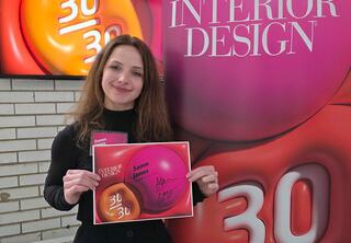 A woman holds a signed certificate while posing in front of a colorful backdrop celebrating her recognition in interior design.