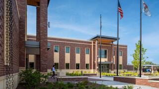 A modern brick building features large windows, a welcoming entrance, and flagpoles, surrounded by landscaped greenery.