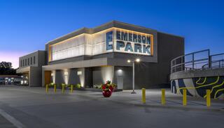 A modern building with a prominent "Emerson Park" sign, featuring bright lights and colorful landscaping, set against a twilight sky.