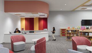 A bright, welcoming reception area features a curved desk, colorful wall panels, cozy seating, and a nearby bookshelf filled with children's books.