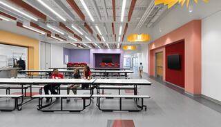 A bright cafeteria features modern design elements, colorful accents, and several long tables, with students seated and engaging.