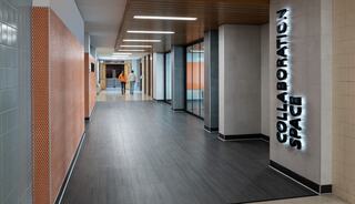 A modern hallway features sleek flooring, orange-accented walls, and illuminated signage for a collaboration space. Two figures walk down the corridor.