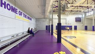 A gymnasium features wooden basketball floors, purple padding along the walls, and bleachers with a few people seated.