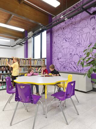 A vibrant library space features children engaged at a round table, surrounded by colorful chairs and a playful mural on the wall.