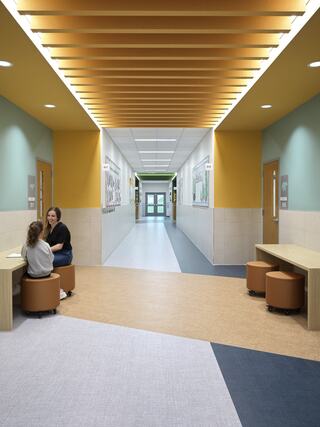 A brightly lit hallway features colorful walls and seating areas, with two people engaged in conversation. Artwork decorates the walls.