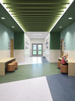 A modern hallway features green acoustic panels, wooden seating areas, and vibrant flooring, creating an inviting educational space.
