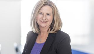 A smiling woman with shoulder-length blonde hair, wearing a black blazer over a purple blouse, poses in a bright, modern setting.
