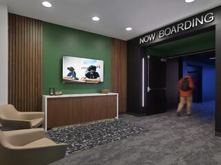 A modern waiting area features a sleek design, a large screen displaying a video, and a dark hallway marked "NOW BOARDING."