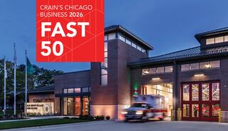 A modern fire station is illuminated at dusk, featuring a red ambulance outside, with a banner announcing Crain's Chicago Business Fast 50.