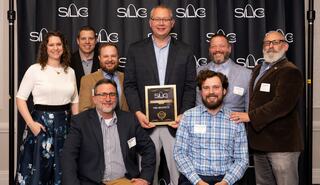 A group of eight individuals poses together, smiling, with one person holding an award in front of a branded backdrop.
