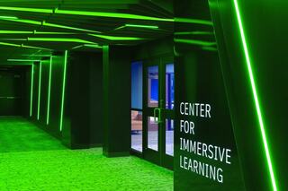 A corridor illuminated with green lighting leads to a door marked "Center for Immersive Learning," showcasing a modern, tech-focused environment.