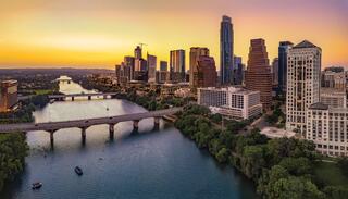 A vibrant city skyline at sunset, reflecting on a river, with lush greenery and a bridge connecting both sides.